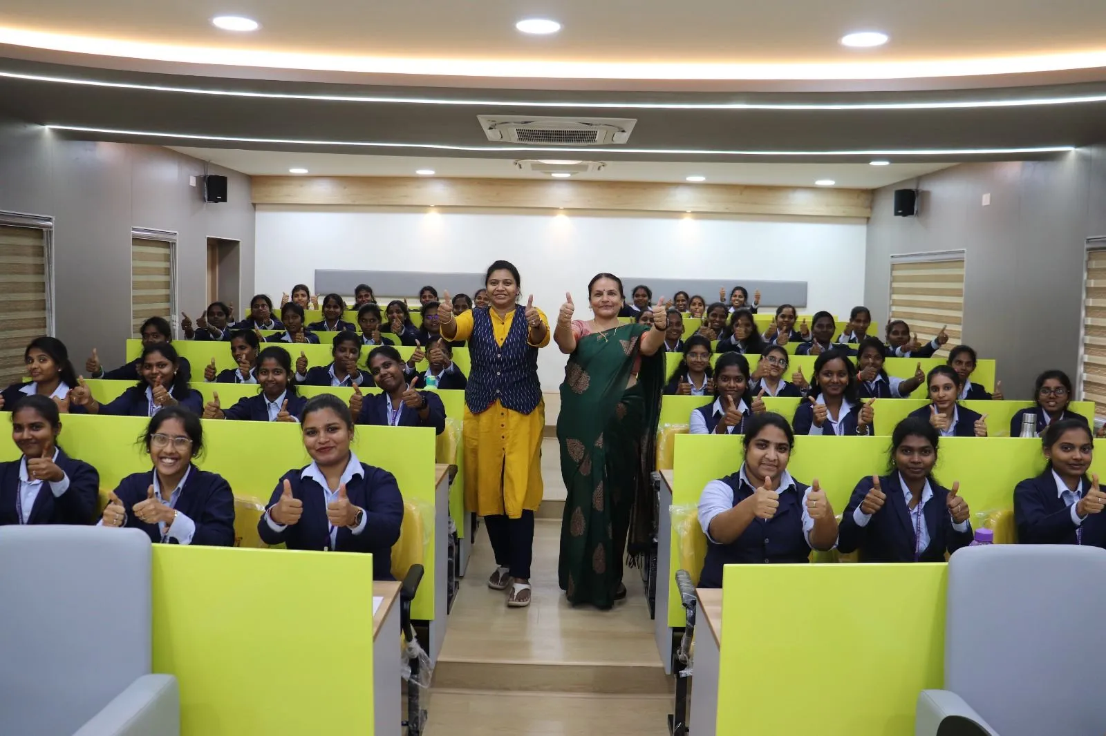 MBA students in navy blazers giving a thumbs-up from their desks in a TIBM lecture hall, with two faculty members standing in the aisle